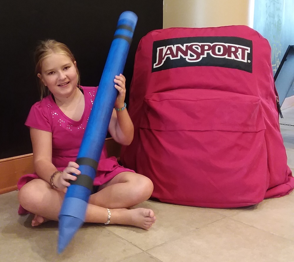 Child holding large crayon and sitting in front of life-size backpack