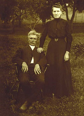 Very old photo of a man in a chair with a woman beside him holding onto the back of the chair